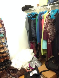 Right side of a walk-in closet with clothes on hangers, with piles of boxes, clothes, boots, and bags on the floor. 
