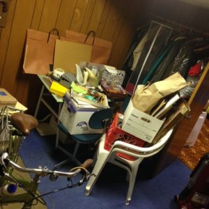A room with piled of arts and papers on a table. Bags and boxes of misc items on top of chairs. Bike handbar is visable in the photo.