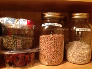 two bali glass jars labeled wheatberry and barley. Plus a plastic container of dried tomatoes on bottom of a clear plastic container or green lentils on a cabinet shelf