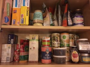 food pantry with cereals on left side of top shelf and bags of grains lined up. Second shelf, below are canned foods, box of pasta, containers of dried mushrooms and jars of honey